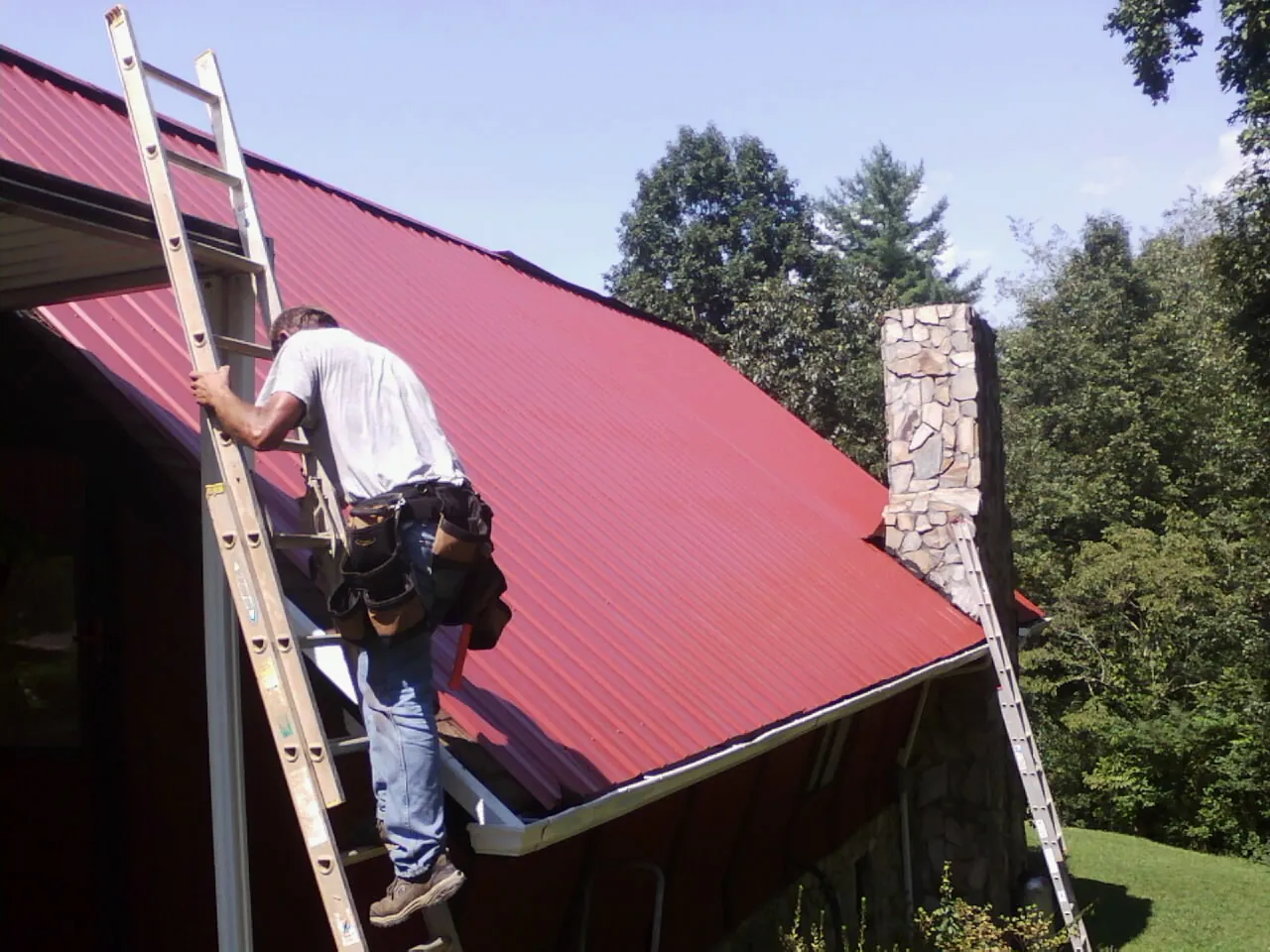 Roofer on a ladder beside a red metal roof and stone chimney — Soffit & Fascia Repair by Roofing Brothers in Beaufort